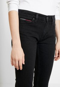 Jeans de mezclilla negra con un corte ajustado, que presentan un acento de bolsillo en rojo, blanco y azul. Cierre con cremallera y botón, textura suave.