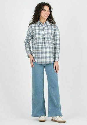 Femme aux cheveux bouclés souriante, portant une chemise à carreaux grise, un jean bleu à jambes larges et des baskets blanches, debout devant un fond uni.