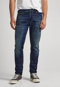 Jeans de mezclilla azul oscuro con corte ajustado, que presentan bolsillos frontales y un lavada sutil, combinados con zapatillas grises y una camiseta blanca.