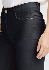 Pantaloni in denim nero con vestibilità slim, dotati di accessori in metallo argentato, tasche laterali e dettagli di cucitura discreti. Il tessuto ha una texture liscia.