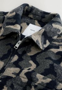 Kraag en ritsdetails van een wollen jas met een grijs, beige en zwart camouflagepatroon en een wit label met de tekst "NEXT."