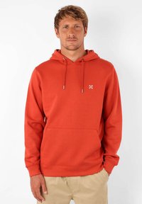 Haut à capuche rouge en tissu doux avec une poche avant, un cordon de serrage pour la capuche et un petit logo blanc sur la poitrine. Il est doté de poignets et d'un ourlet côtelés.