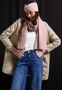 Veste matelassée beige clair, chemise blanche, jeans en denim et écharpe en tricot rose à texture côtelée. Bandeau tricoté rose et gants complètent la tenue.