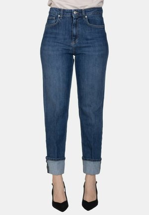 Donna che indossa jeans blu scuro con risvolti e tacchi neri a punta, in piedi davanti a uno sfondo chiaro e semplice.