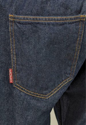 Dunkelblauer Denim-Stoff mit auffälligen orangefarbenen Nähten. Verfügt über eine Gesäßtasche mit einem roten "DADA"-Label. Strukturiert Oberfläche, lässiges Design.