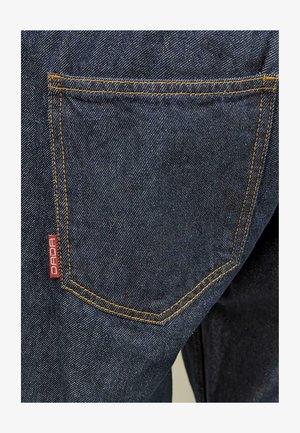 Dunkelblauer Denim-Stoff mit auffälligen orangefarbenen Nähten. Verfügt über eine Gesäßtasche mit einem roten "DADA"-Label. Strukturiert Oberfläche, lässiges Design.