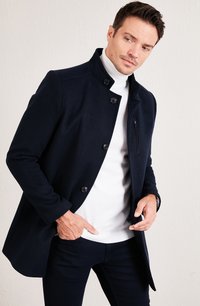 Cappotto blu navy in misto lana con colletto rialzato, chiusure a bottoni, tasca sul petto con zip e tasche laterali; indossato su un dolcevita bianco.