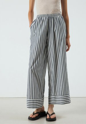 Volwassene draagt losse broek met brede pijpen en verticale zwart-witte strepen en zwarte platformsandalen, staand tegen een effen achtergrond.