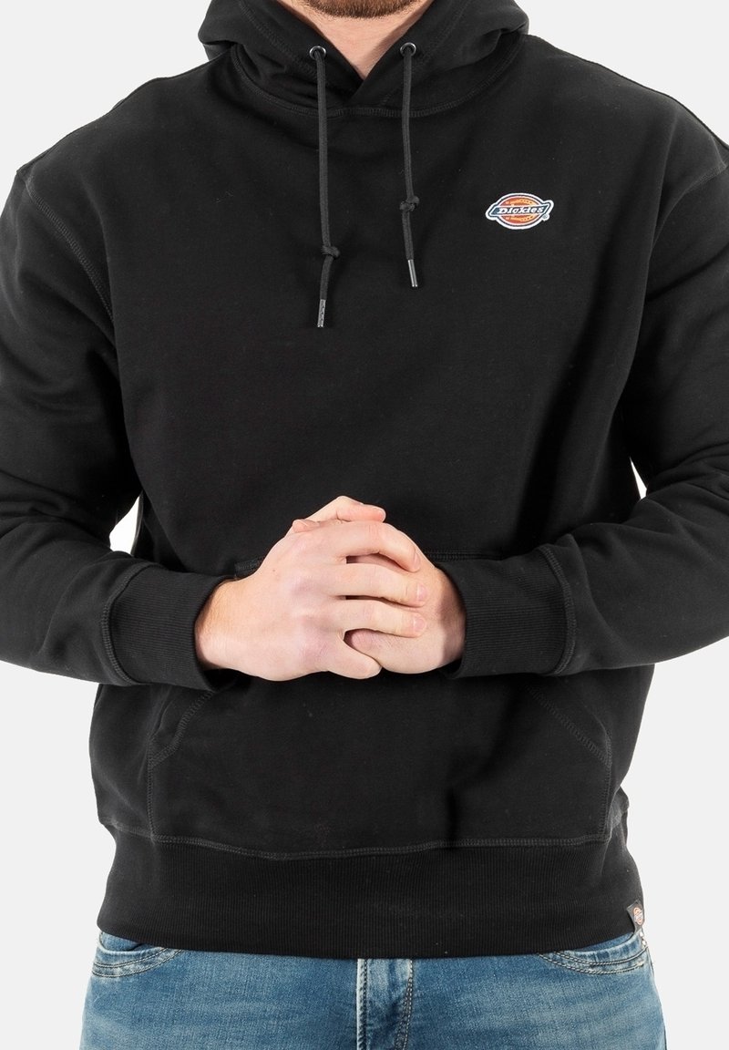 Svart hoodie av mjukt tyg med en kenguruficka, justerbara dragskor och en liten logga på bröstet. Bekväm passform och design.