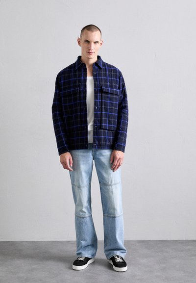 Navy and black checkered flannel shirt, featuring two front pockets, paired with light blue jeans and black sneakers. Smooth texture and relaxed fit.