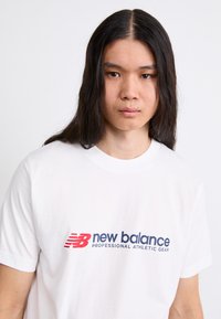 Camiseta de algodón blanca con un logo gráfico en rojo y azul marino que dice "new balance PROFESSIONAL ATHLETIC GEAR" en el pecho.