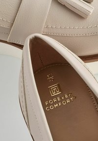 Beiget värikkäät loafersit, joissa on nahkatuntu, koristeelliset tupsut ja kirjonnat. Sisäpuolella näkyy kultainen logo ja kokomerkinnät.
