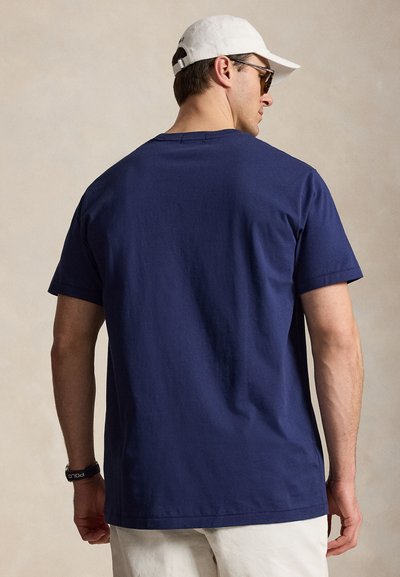 Man wearing navy blue short-sleeve t-shirt, white cap, sunglasses, beige pants, and black wristwatch, facing away from the camera.