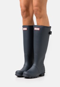 Botas de agua de goma negras hasta la rodilla con suela texturizada, con acentos del logo "HUNTER" en rojo y blanco en la parte delantera.