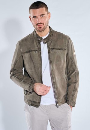 Man with short hair wearing a gray suede jacket over a white t-shirt, standing with one hand adjusting the jacket zipper.