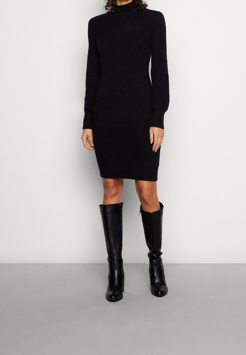 Femme portant une robe en maille noire ajustée à manches longues avec un col montant et des bottes noires montant aux genoux, se tenant devant un fond clair.