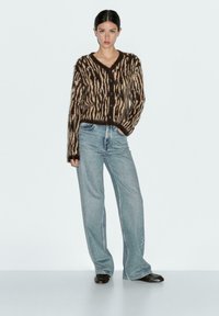 Cardigan a righe marroni e beige con bottoni, abbinato a un paio di jeans a gamba larga blu e scarpe scure, su uno sfondo chiaro.