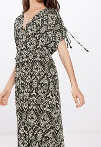 Langes, locker sitzendes Kleid in Dunkelgrün mit weißen geometrischen Mustern. Es verfügt über einen tiefen V-Ausschnitt und geraffte Ärmel mit Bindebändern.