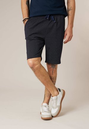 Homme portant un short bleu marine texturé avec cordon de serrage et des baskets blanches, debout avec une jambe croisée devant l'autre, montrant des tatouages sur les jambes.