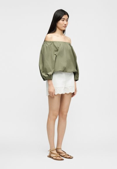 Γυναίκα με πράσινο ελιάς μπλούζα έξω από τους ώμους, λευκά σορτς με δαντέλα στο τελείωμα και καφέ παντόφλες με επίπεδη σόλα, στέκεται μπροστά σε απλό φόντο.