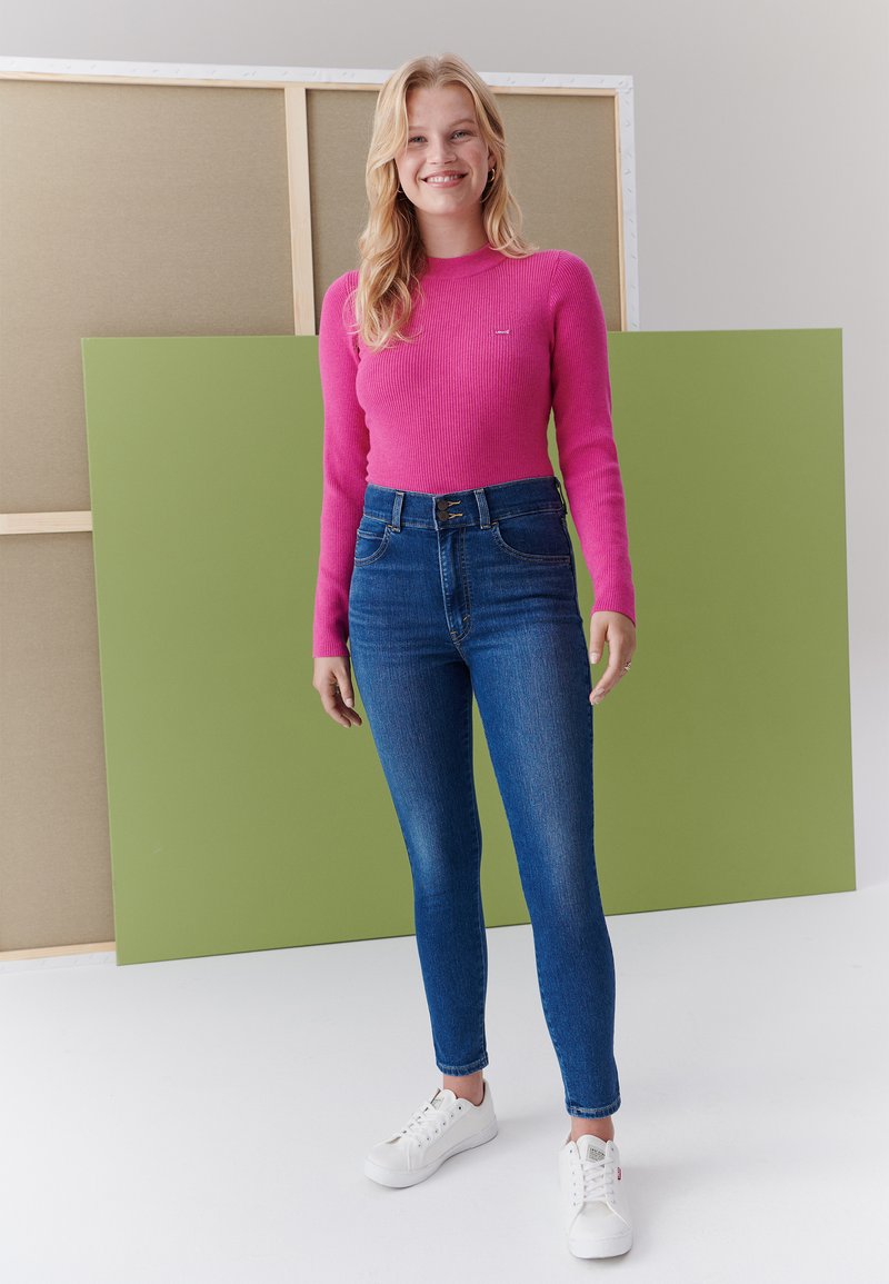 Top fucsia a maniche lunghe a coste, jeans skinny blu a vita alta e sneakers bianche con lacci, contro sfondi verdi e beige.