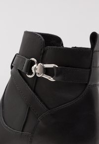 Black leather ankle boot with a smooth texture, featuring a decorative silver buckle and strap detail on the upper part.