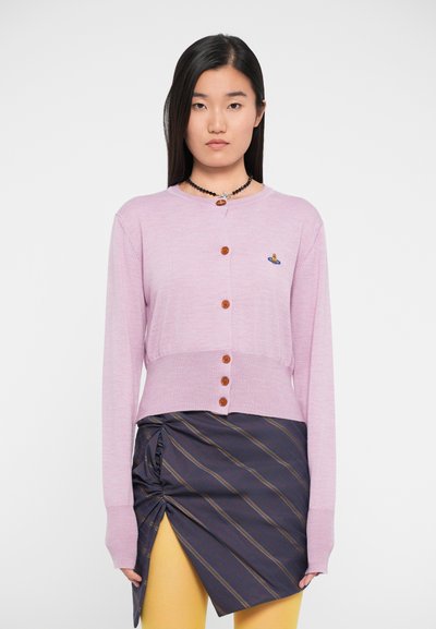 Cardigan rose clair avec une taille ajustée, des boutons en bois et un petit détail brodé sur la poitrine. Le modèle porte une tenue superposée.