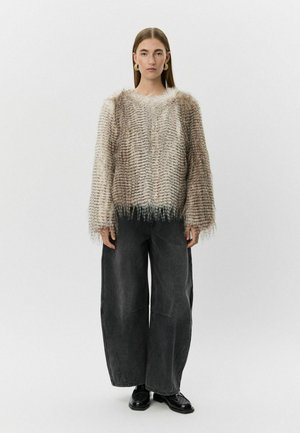 Textúrált, világos bézs pulóver, rojtos aljjal, laza fekete farmernadrág felett viselve. Nagy, kötött mintázat vízszintes vonalakkal.