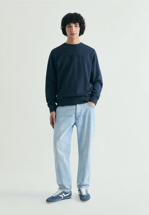 Ung mand med krøllet hår iført en marineblå sweatshirt, lyseblå jeans og marineblå sneakers, stående med den ene hånd i lommen.