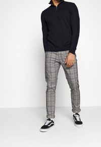 Zwarte gebreide pullover met een kwart ritssluiting, gecombineerd met grijs geruite slim fit broek en zwarte sneakers met witte zolen.
