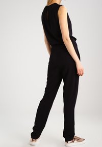 Svart ärmlös topp med draperad rygg, kombinerad med svarta smala byxor och metalliska sneakers, vilket skapar en strömlinjeformad, monokromatisk look.