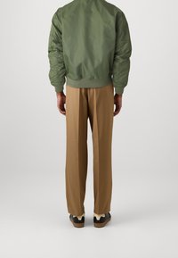 Zelená bomber bunda s žebrovanými manžetami, doplněná béžovými širokými kalhotami a černými teniskami, pohled zezadu na světlém pozadí.