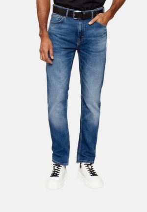 Hombre que lleva puestos unos vaqueros ajustados azules con cinturón negro y zapatillas blancas, de pie contra un fondo liso.