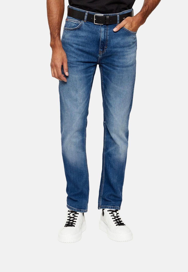 Homme portant un jean slim bleu avec une ceinture noire et des baskets blanches, debout devant un fond uni.