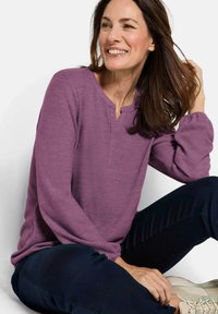 Lila Langarmshirt mit V-Ausschnitt und gerafften Ärmeln, kombiniert mit dunklen Hosen und hellen Turnschuhen. Lässig, weicher Stoff und bequeme Passform.