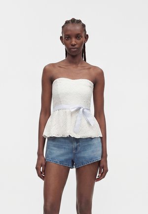 Vrouw met gevlochten haar die een witte kanten strapless top met een taallint en blauwe denimshorts draagt, staand tegen een effen achtergrond.