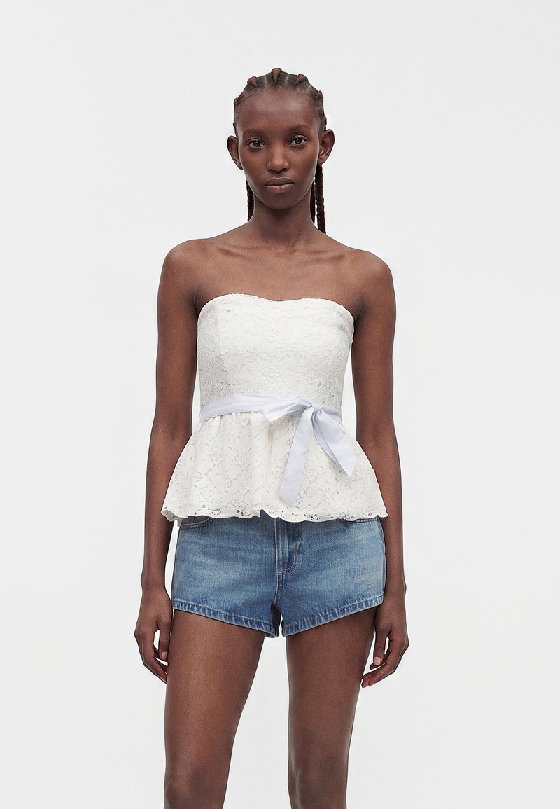 Femme aux cheveux tressés portant un haut en dentelle blanc sans bretelles avec un ruban à la taille et un short en jean bleu, debout devant un fond uni.
