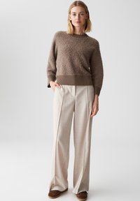 Brauner, flauschiger Pullover mit geripptem Saum, kombiniert mit hellbeigen, weit geschnittenen Hosen. Das Model trägt braune Schuhe und steht vor einem neutralen Hintergrund.