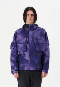 Purple patterned jacket featuring multiple front pockets, adjustable hood, and drawstrings. Made from lightweight, textured fabric.