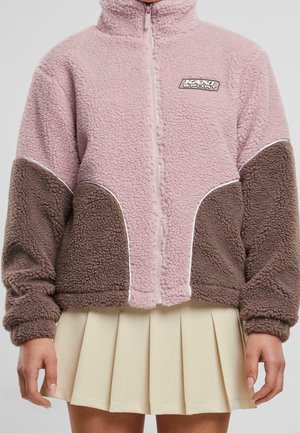 Light pink and brown fleece jacket with zipper and logo on left chest, worn with a beige pleated skirt.