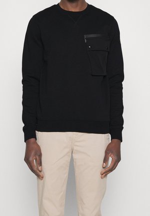 Homme portant une chemise noire à manches longues avec une grande poche boutonnée sur la poitrine gauche et un pantalon beige, debout sur fond blanc.