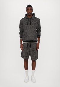 Černá mikina s kapucí a krátké kalhoty s geometrickým vzorem, šňůrková kapuce a žebrované manžety. Nosič má na sobě bílé tenisky a kotníkové ponožky.