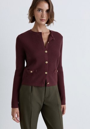 Frau trägt eine gerippte burgunderrote Strickjacke mit goldenen Knöpfen und olivgrünen, hoch taillierten Hosen vor einem einfarbigen hellen Hintergrund.