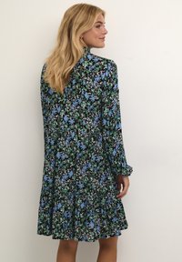 Schwarz gekleidetes Kleid mit floralem Muster in Blau und Grün. Lange Ärmel, taillierter Oberteil, ausgestellter Rock mit Rüsche am Saum. Glatte Stofftextur.