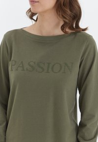 Olivovozelená dlhá košeľa s dlhými rukávmi a okrúhlym výstrihom, na ktorej je v textúrovanom tónovom vyšívaní napísané slovo "PASSION". Vyrobená z jemnej bavlny.