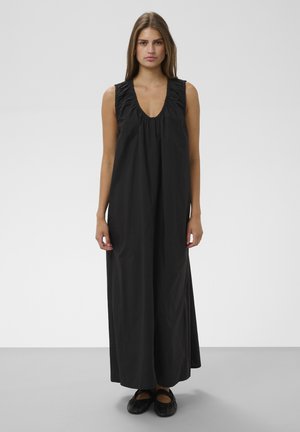 Frau steht, trägt ein ärmelloses schwarzes Maxikleid mit gerafftem Ausschnitt und schwarzen flachen Schuhen vor einem schlichten hellen Hintergrund.