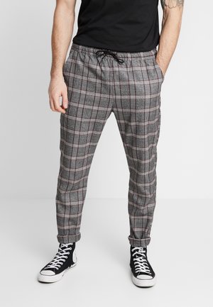 Mann trägt graue karierte Jogginghose mit Kordelzug und umgeschlagenen Bündchen, schwarze High-Top-Sneaker und ein schwarzes T-Shirt, steht mit den Händen in den Taschen.