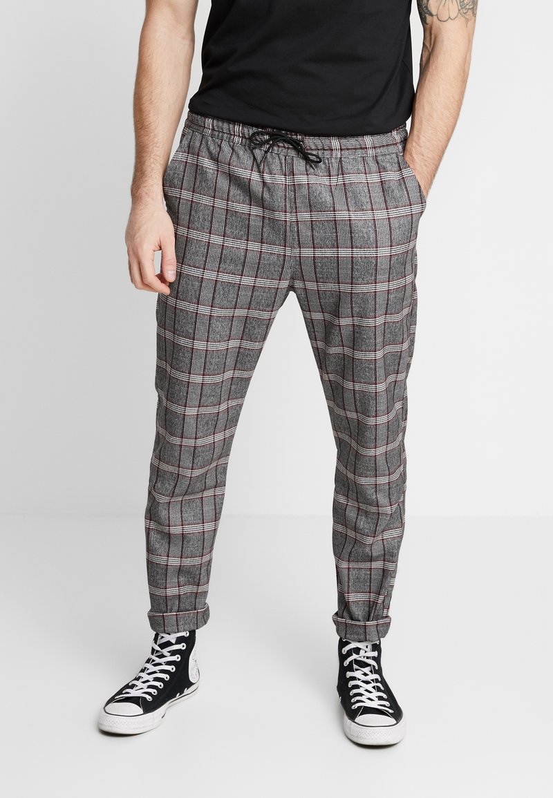 Homme portant un pantalon à carreaux gris avec cordon de serrage et revers roulés, des baskets montantes noires et un t-shirt noir, debout les mains dans les poches.