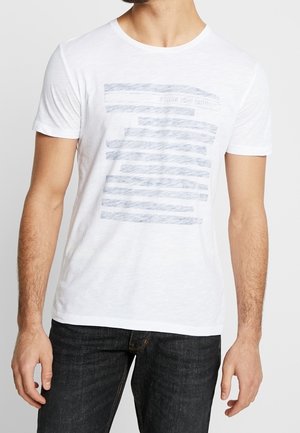 Man wearing a white short-sleeve T-shirt with faded horizontal blue stripes and dark jeans, standing against a plain background.