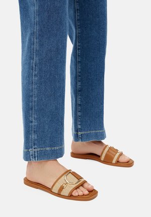 Brązowe i beżowe klapki na płaskiej gumowej podeszwie z plecionymi paskami tekstylnymi, okrągłym metalowym akcentem, zestawione z niebieskimi dżinsowymi spodniami.